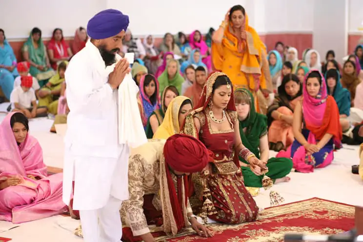 Groom wearing pagri on hands and knees in front of holy book